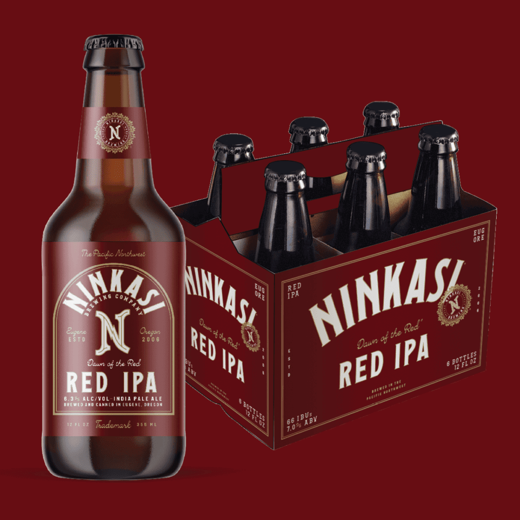 A Ninkasi "Dawn of the Red" Red IPA bottle and 6-pack box against a deep red background. The amber-hued bottle showcases the Ninkasi logo at the top, with "Dawn of the Red" and "Red IPA" prominently displayed below. The label also indicates a 6.3% alcohol volume and that it's brewed in Eugene, Oregon. The accompanying box, holding six identical bottles, mirrors the bottle's design, highlighting the "Red IPA" and the Ninkasi branding.