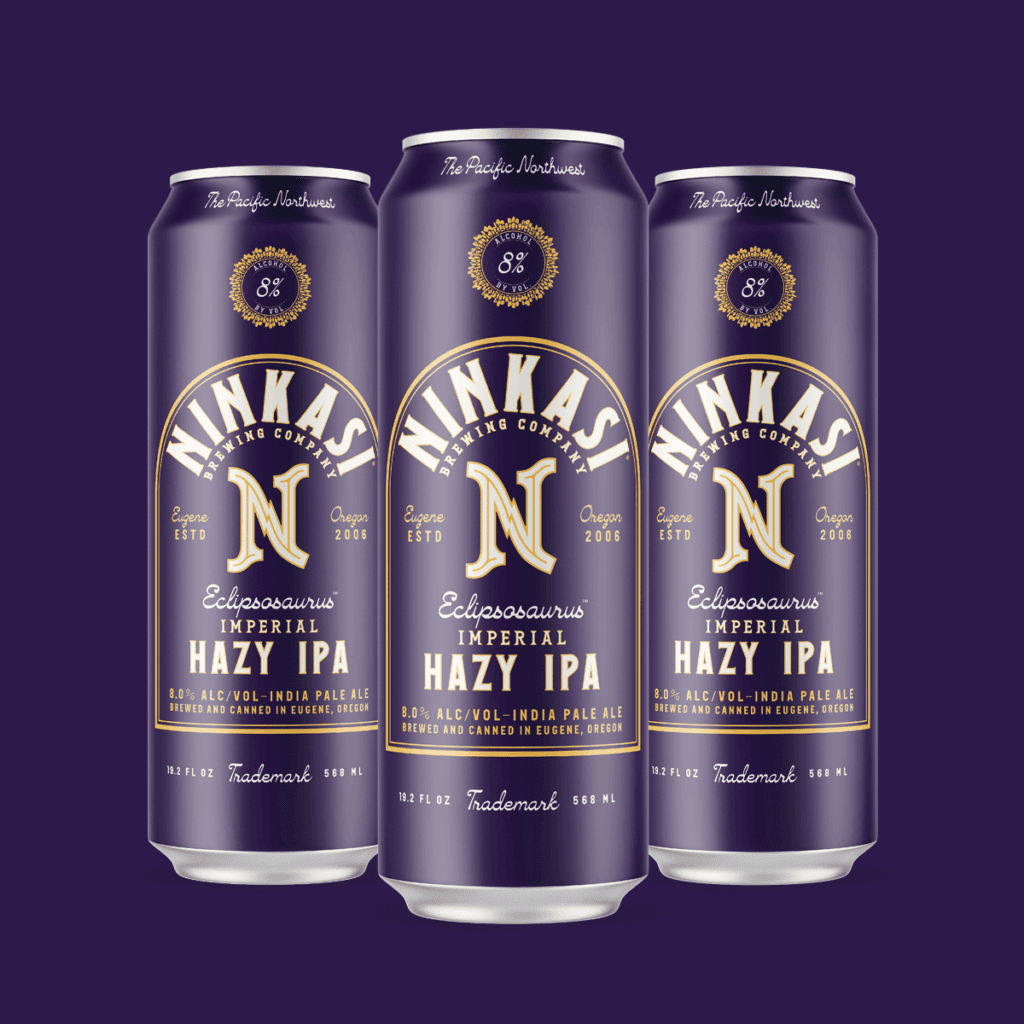 Three tall cans of Ninkasi's "Eclipsosaurus Imperial Hazy IPA" against a deep purple background. The cans sport a rich purple and gold design, featuring the label "The Pacific Northwest". Each can has an emblem indicating "8% ALC/VOL", representing its alcohol content. They are described as "Imperial Hazy IPA" and are brewed and canned in Eugene, Oregon, with a founding date of 2006 indicated on each can. The Ninkasi Brewing Company logo is displayed prominently, and each can specifies a volume of 19.2 fl oz.