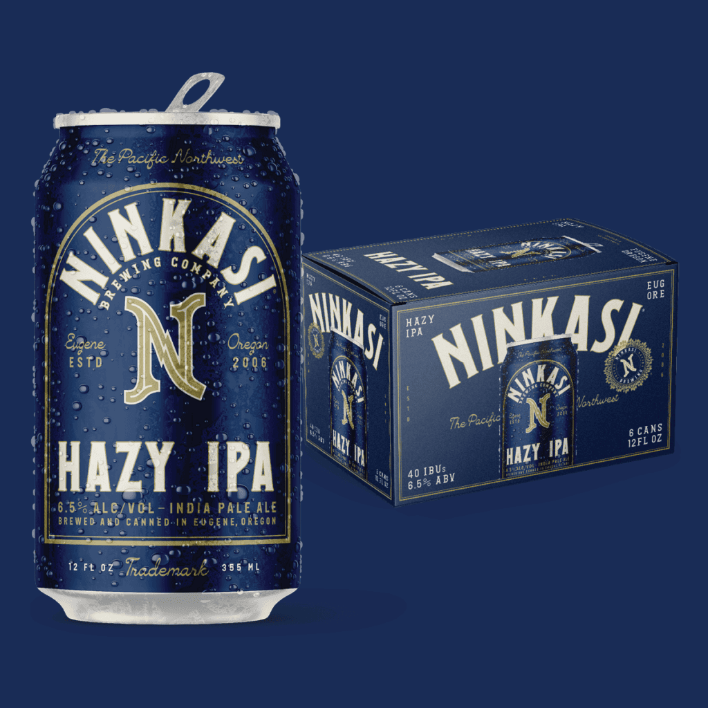 A can and a 6-pack box of Ninkasi Hazy IPA beer. The can and box have a deep blue background with elegant gold and white lettering. Prominently featured is the Ninkasi Brewing Company logo, which is a stylized 'N' with a wing-like design, and the words "HAZY IPA" in bold lettering. Below, it reads "6.5% ALC/VOL - INDIA PALE ALE" and "BREWED AND CANNED IN EUGENE, OREGON." Water droplets are visible on the can, suggesting it's cold. The box repeats the design elements of the can and indicates it contains 6 cans, each with 12 fl oz. The overall look is premium and crafted, highlighting the Pacific Northwest origin of the beer.